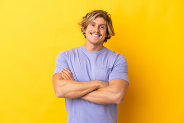 Handsome blonde man isolated on yellow background keeping the arms crossed in frontal position