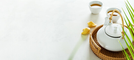 Asian tea concept, two white cups of tea and teapot surrounded with green dry tea  with space for a text on light background. Wide banner. - obrazy, fototapety, plakaty