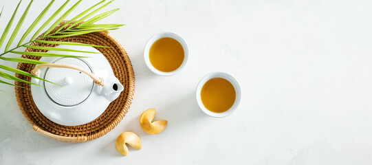 View from above of Asian tea concept, two white cups of tea, fortune cookies and teapot surrounded with dry green tea on light background.Wide banner. - obrazy, fototapety, plakaty