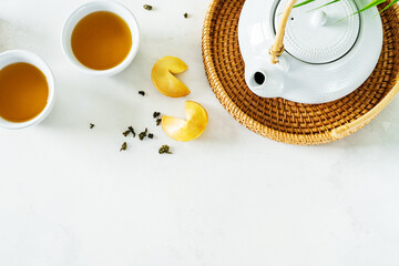 View from above of Asian tea concept, two white cups of tea, fortune cookies and teapot surrounded with dry green tea on light background