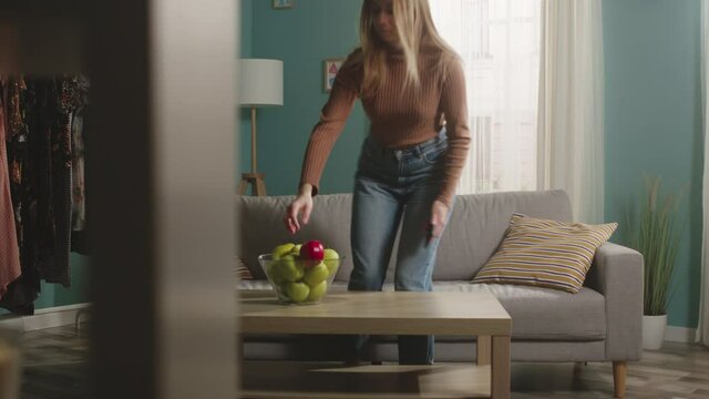 The girl in jeans, a brown sweater enters the room with a mobile phone, takes a red apple from the table and lies down on the sofa