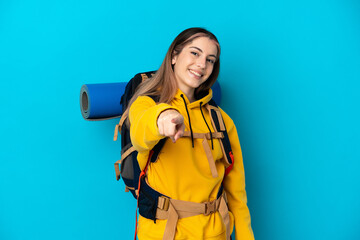 Young mountaineer woman with a big backpack isolated on blue background pointing front with happy expression