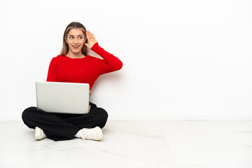 Young caucasian woman with a laptop sitting on the floor listening to something by putting hand on...