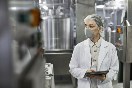 Waist up portrait of young woman wearing mask and holding digital tablet during quality control inspection at food factory, copy space