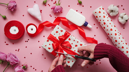 Step 12.Step-by-step instructions for wrapping gifts for Valentine's Day. Woman wraps a gift with wrapping white paper in heart and seals the edges with glue on a pink background top view, flat lay
