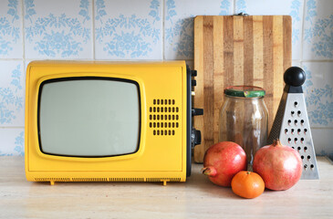 An old yellow vintage TV stands in the kitchen next to fruit and kitchen utensils against a...