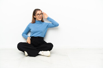 Young Caucasian woman sitting on the floor listening to something by putting hand on the ear