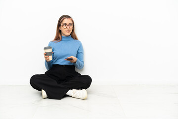 Young Caucasian woman sitting on the floor holding coffee to take away and a mobile while thinking something