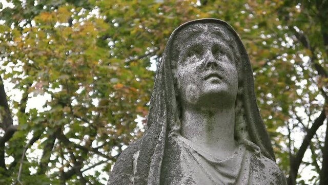 Stone sculpture of woman in cemetery. Madonna figure with defocused leaves in the background.