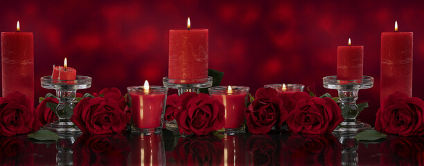 Fototapeta na wymiar Lit red candles in transparent candlesticks illuminate roses on a mirror surface on a red background. Bokeh in shape of a heart. Valentine's day or romantic evening invitation