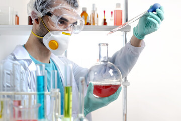 Young scientist in protective equipment holding a round flask looking at a drop of substance from a...