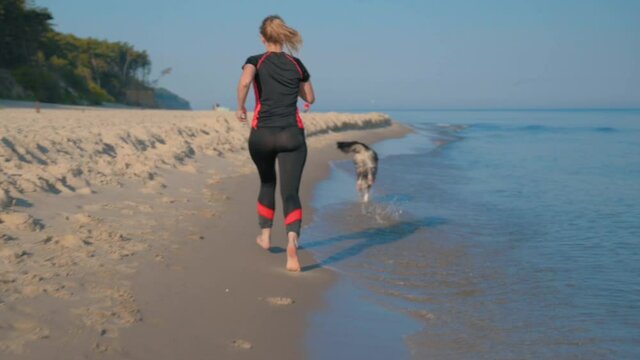 Beautiful active sport girl walking with Australian Shepherd dog on the beach. Woman runs and plays with Aussie dog. Female owner with her happy pet near summer sea. Back view.