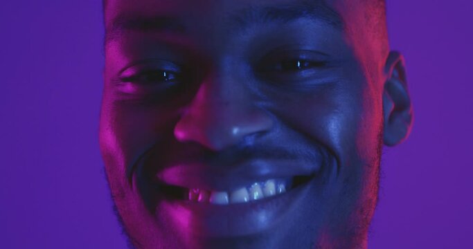 Young positive african american man turning face to camera and widely smiling, neon lights background