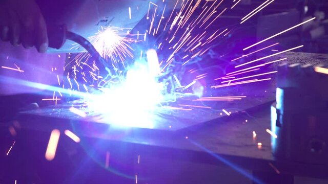 Close-up blacksmith welder in protective mask works with metal steel and iron using a welding machine, bright sparks and flashes in extreme slow motion