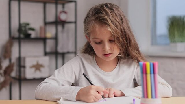 Cute little girl are drawing pictures with felt tip pen on the white paper album sitting at the table. Playing alone, creative art activity at home. Spending free time at home during school holidays