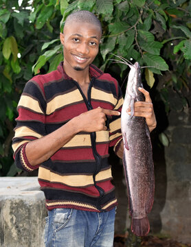 Guy Holding A Big Cat Fish Up High In His Hands With Joyful Expression On His Face. A Nigerian Cat Fish Farmer 