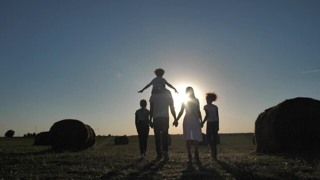 Silhoutte of multinational family, african american man, caucasian wife and two mixed race preteen daughters enjoying walk hand in hand on wheat field, youngest girl riding on dad's shoulders