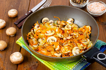 Frying pan with fried mushrooms champignons, onions and carrots on wooden background. Studio Photo