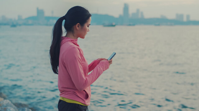 Portrait of a sportswoman using smartphone checking social media feeds during taking rest after running at the seaside. Sportswomen use cellphones to send messages to friends while standing resting.