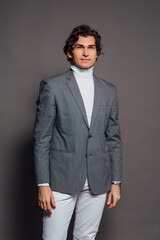 Fototapeta premium Tall handsome man dressed in white turtleneck, white jeans and grey jacket posing on the grey background
