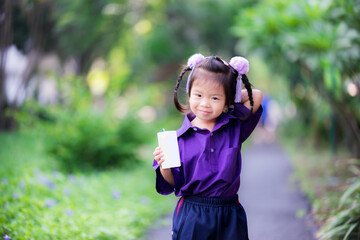 Pupil with milk cartons for morning walk in the park near school. Children drink boxed milk. Child smiled sweetly, showing little embarrassment. Asian kid wearing purple sports shirt. Girl 3 years old