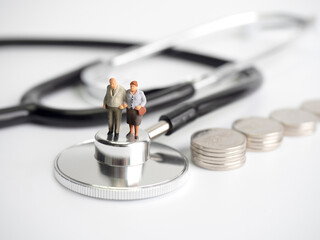 Miniature old people standing on medical stethoscope with coins stack on white background,...