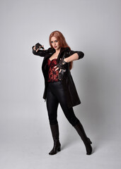 full length portrait of girl with long red hair wearing dark leather coat, corset and boots. Standing pose facing front on with  magical hand gestures against a  studio background.
