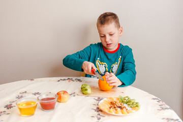 a nine-year-old boy sits at a table and cuts an orange. The child prepares his own breakfast at home