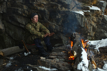 Extreme traveller warms his hands near the bonfire, hiding from the wind under the overhanging cliffs in cold weather.