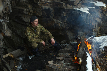 Tired traveller warms up near a campfire divorced next to an overhanging cliff protecting him from the wind.