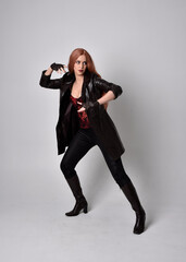 full length portrait of girl with long red hair wearing dark leather coat, corset and boots. Standing pose facing front on with  magical hand gestures against a  studio background.