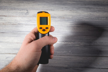 Fototapeta premium A man holds a yellow-black pyrometer on a wooden background. A device for non-contact temperature measurement.