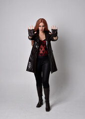 full length portrait of girl with long red hair wearing dark leather coat, corset and boots. Standing pose facing front on with  magical hand gestures against a  studio background.