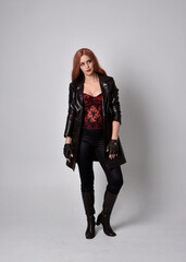 Fototapeta na wymiar full length portrait of girl with long red hair wearing dark leather coat, corset and boots. Standing pose facing front on with magical hand gestures against a studio background.
