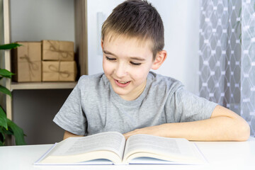 A boy reads a book. A schoolboy in a gray T-shirt at home at a white table looks into a book.