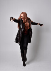 full length portrait of girl with long red hair wearing dark leather coat, corset and boots. Standing pose facing front on with  magical hand gestures against a  studio background.