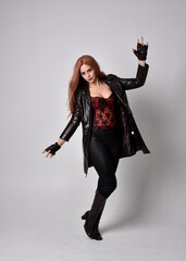 full length portrait of girl with long red hair wearing dark leather coat, corset and boots. Standing pose facing front on with  magical hand gestures against a  studio background.