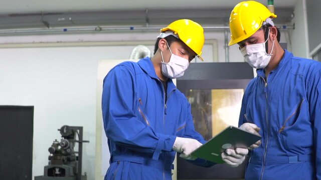 Asian male worker in safety uniform wearing protective mask using digital tablet working with colleague at factory industrial construction