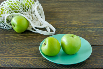 Organic Granny Smith apples on a plate with mesh textile bag on wooden table.