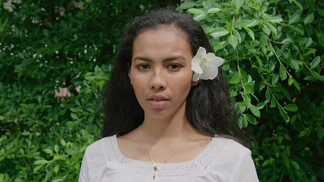 Portrait of a young beautiful Thai woman with a flower in her hair standing in a garden and looking straight into the camera on a sunny day in slow motion.