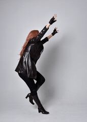 full length portrait of girl with long red hair wearing dark leather coat, corset and boots. Standing pose facing back view with  magical hand gestures against a  studio background.