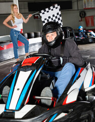 Diligent man in helmet driving car for karting in sport club, friendly smiling woman with flag on background