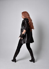 full length portrait of girl with long red hair wearing dark leather coat, corset and boots. Standing pose facing front on with  magical hand gestures against a  studio background, low camera angle.