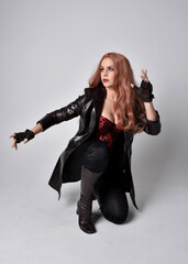 full length portrait of girl with long red hair wearing dark leather coat, corset and boots. Sitting pose facing front on with  magical hand gestures against a studio background.