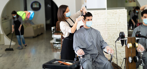 Female hairdresser making hair styling for young man in protective mask and gloves, working day...