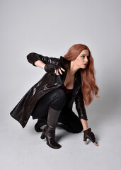 full length portrait of girl with long red hair wearing dark leather coat, corset and boots. Sitting pose facing front on with  magical hand gestures against a studio background.