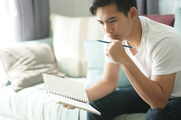 thoughtful asian young student man teenager thinking holding notebook on sofa at home