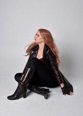 full length portrait of girl with long red hair wearing dark leather coat, corset and boots. Sitting pose facing front on with  magical hand gestures against a studio background.