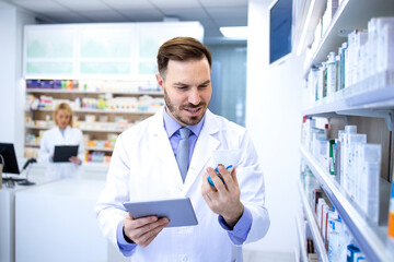 Handsome male pharmacist in white coat holding medicine in pharmacy store or drugstore and checking price on tablet computer. Healthcare and apothecary.