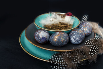 Easter concept of coloring eggs in hibiscus, eco dyes. Front view, black background and turquoise plates. Bird in nest on background. Feathers as decoration.